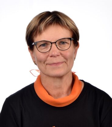 Henkilö, jolla on lyhyet hiukset ja silmälasit, on yllään oranssi huivi ja musta paita ja hymyilee valkoista taustaa vasten.