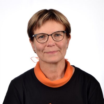 Henkilö, jolla on lyhyet hiukset ja silmälasit, on yllään oranssi huivi ja musta paita ja hymyilee valkoista taustaa vasten.