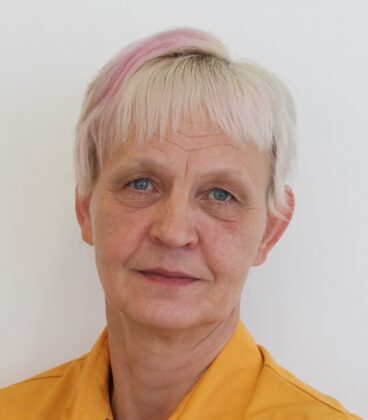 Vanhempi henkilö, jolla on lyhyet valkoiset hiukset ja aavistuksen vaaleanpunainen, yllään oranssi paita ja katsoo suoraan kameraan neutraalilla ilmeellä tavallista valkoista taustaa vasten.