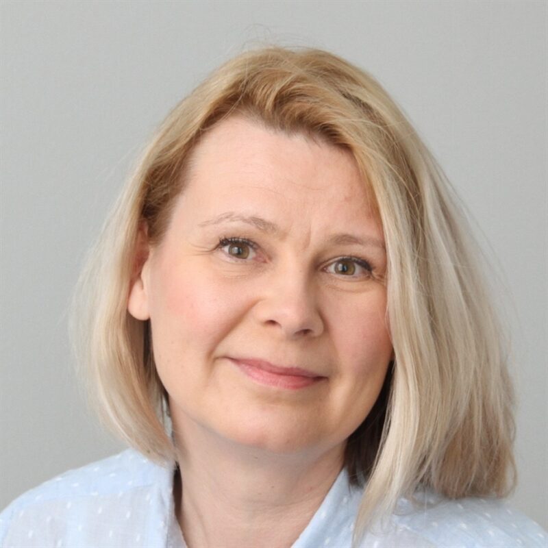 Ihminen, jolla on olkapäille ulottuvat vaaleat hiukset, hymyilee lempeästi. Heillä on yllään vaaleansininen paita ja he asettuvat harmaata taustaa vasten.