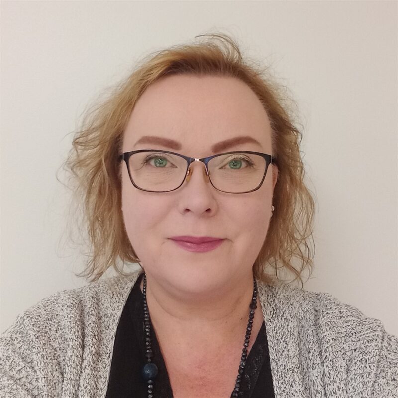 Kuvassa valkoista taustaa vasten aaltoilevat, vaaleanruskeat hiukset, silmälasit ja neutraali ilme. Heillä on yllään harmaakuvioinen villapaita mustan topin päällä ja helmillä koristeltu kaulakoru.