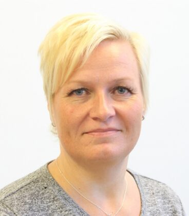 Henkilö, jolla on lyhyet vaaleat hiukset ja neutraali ilme, on kameraa päin. Heillä on yllään harmaa toppi ja herkkä kaulakoru. Tausta on tavallinen valkoinen.