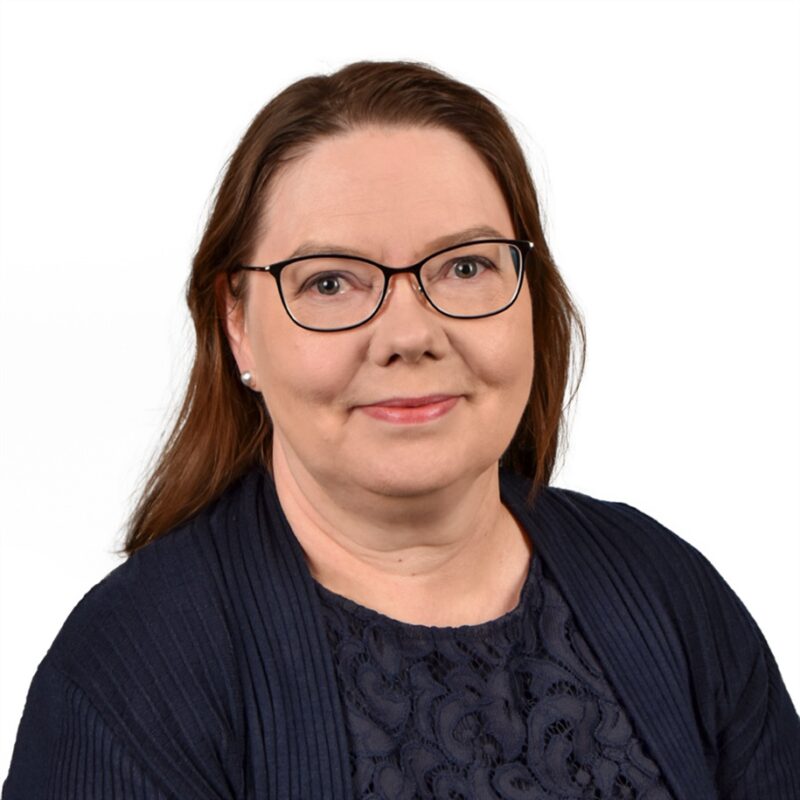 Nainen, jolla on pitkät ruskeat hiukset, yllään silmälasit, tummansininen pusero ja tummansininen kuviollinen toppi. Hän hymyilee pehmeästi ja tausta on pelkkä valkoinen.