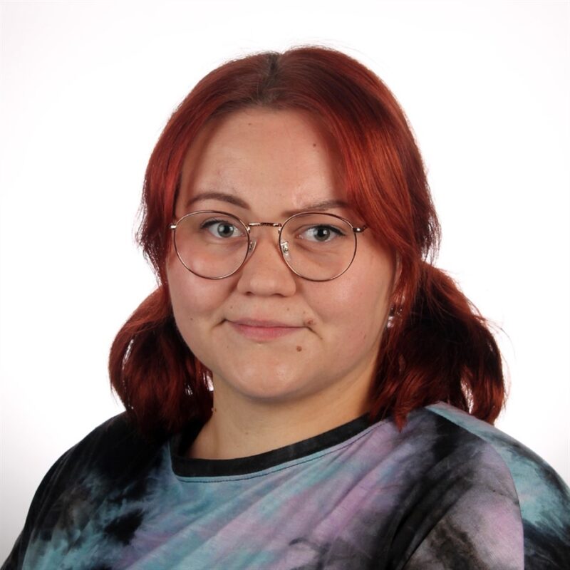 Hartioihin ulottuvat punaiset hiukset ja silmälasit, solmittava paita yllään, hymyilee lempeästi kameralle tavallista valkoista taustaa vasten.