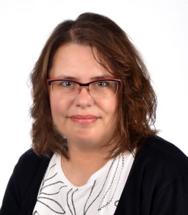Hartioihin ulottuvat ruskeat hiukset ja silmälasit omaava henkilö hymyilee hieman kameralle. Heillä on yllään valkoinen paita mustilla kuvioilla ja musta neuletakki tavallista valkoista taustaa vasten.