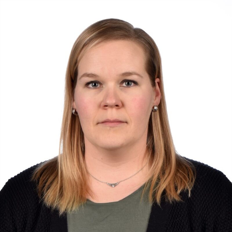 Muotokuva henkilöstä, jolla on olkapäille ulottuvat vaaleat hiukset, yllään tumma pusero vihreän topin päällä, valkoista taustaa vasten.