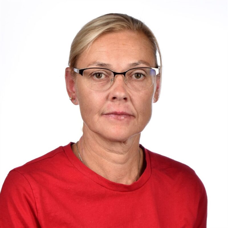 Silmälaseja ja punaista paitaa käyttävä henkilö poseeraa valkoisen taustan edessä. Heillä on lyhyet, vaaleat hiukset ja neutraali ilme.