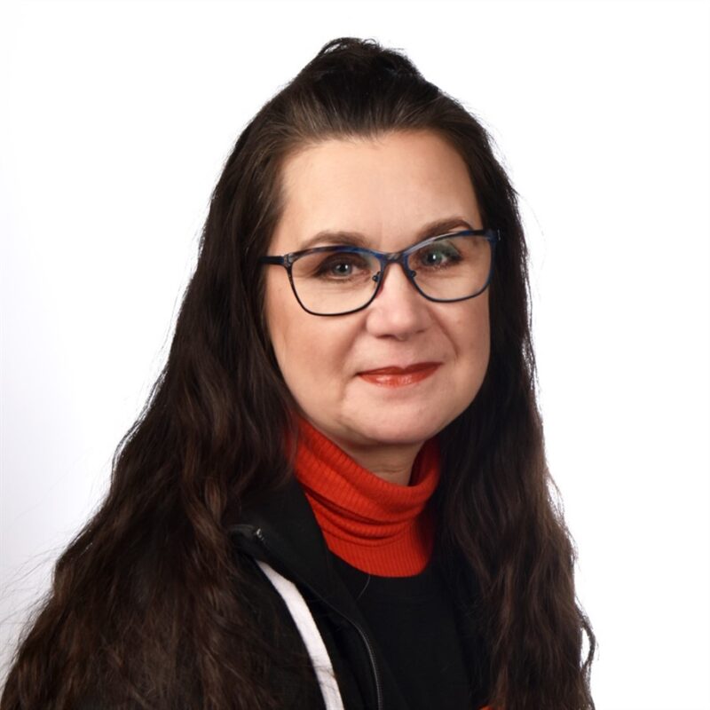 Nainen, jolla on pitkät tummat hiukset ja silmälasit, yllään punainen villapaita ja musta takki, hymyilee kameralle valkoista taustaa vasten.
