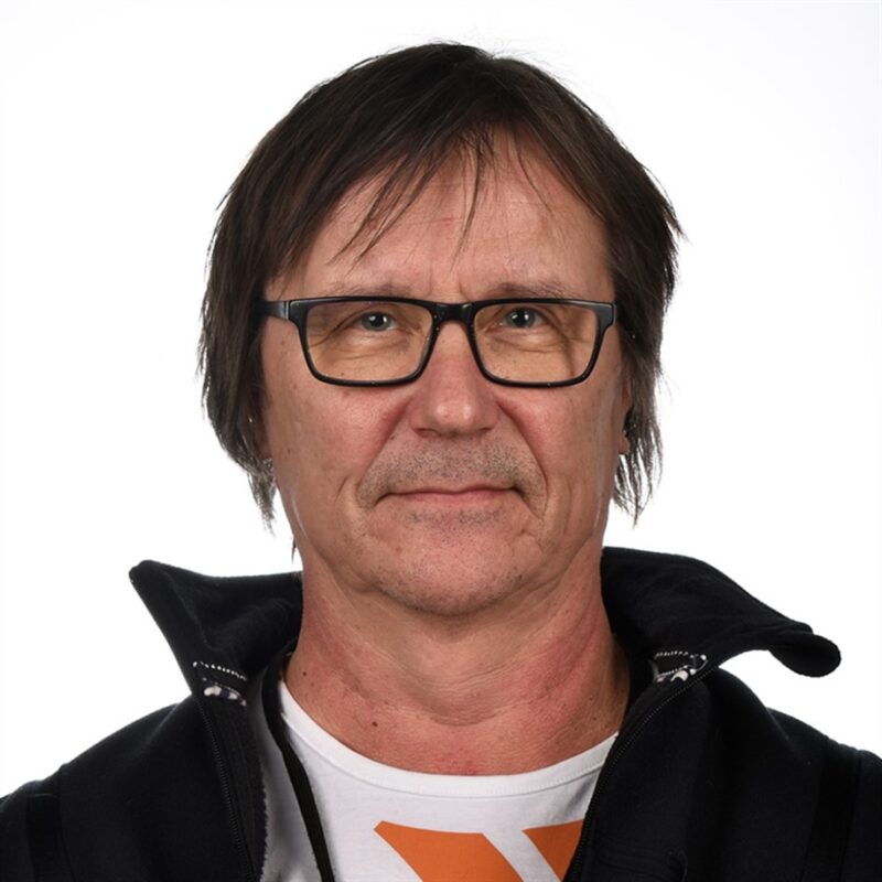 Henkilö, jolla on silmälasit, musta takki ja valkoinen paita, jossa on oransseja yksityiskohtia, katsoo suoraan kameraan. Tausta on tavallinen valkoinen.