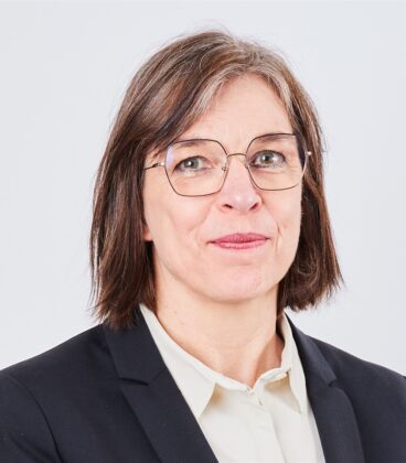 Nainen, jolla on olkapäitä ulottuvat ruskeat hiukset, yllään silmälasit, valkoinen paita ja musta bleiseri, seisoo valkoista taustaa vasten. Hän katsoo kameraa päin hieman hymyillen.