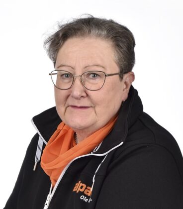 Henkilö, jolla on silmälasit, oranssi huivi ja musta logollinen takki, katsoo kameraan valkoista taustaa vasten.