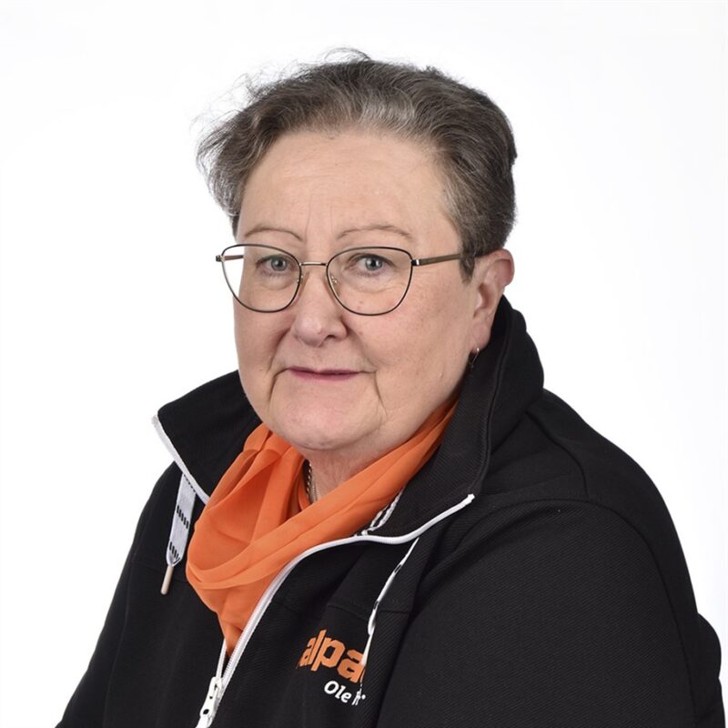Henkilö, jolla on silmälasit, oranssi huivi ja musta logollinen takki, katsoo kameraan valkoista taustaa vasten.