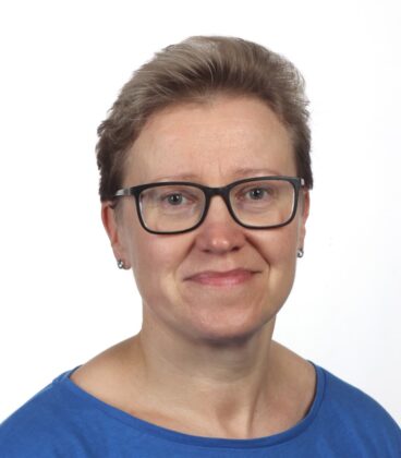 Lyhythiuksinen vaaleanruskea tukka, mustasankaiset silmälasit, pienet korvakorut ja sininen toppi hymyilevät lempeästi kameralle yksinkertaista valkoista taustaa vasten.