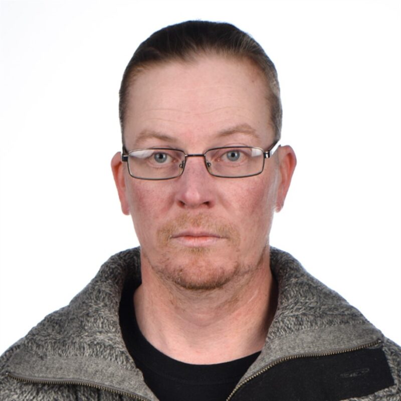 Miehellä, jolla on silmälasit ja lyhyt kampaus, on yllään musta paita ja harmaa vetoketjullinen takki. Hän katsoo suoraan kameraan tavallista valkoista taustaa vasten.