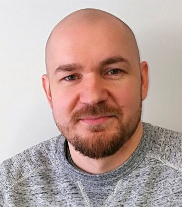 Kalju mies, jolla on parta, hymyilee hieman, yllään vaaleanharmaa neulepusero ja seisoo valkoista taustaa vasten.