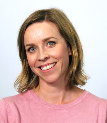 Hymyilevä henkilö, jolla on olkapäille ulottuvat vaaleanruskeat hiukset ja yllään vaaleanpunainen toppi tavallista valkoista taustaa vasten.