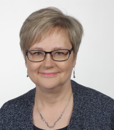 Nainen, jolla on lyhyet, vaaleanruskeat hiukset ja silmälasit, hymyilee kameralle. Hänellä on yllään sininen kuviollinen neuletakki, tumma toppi, pienet korvakorut ja herkkä hopeakaulakoru. Tausta on tavallinen valkoinen.