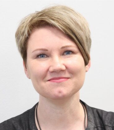 Lyhythiuksinen, vaaleanruskeahiuksinen ja sinisilmäinen nainen hymyilee kameralle. Hänellä on yllään musta paita ja kaulakoru, ja hänen takanaan on yksivärinen vaalea tausta.