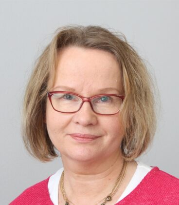 Hartioihin ulottuvat vaaleanruskeat hiukset ja silmälasit omaava henkilö on pukeutunut punaiseen toppiin, jossa on valkoinen kaulus. Niissä on neutraali ilme ja kaulakoru. Taustalla on tavallinen vaaleanharmaa seinä.