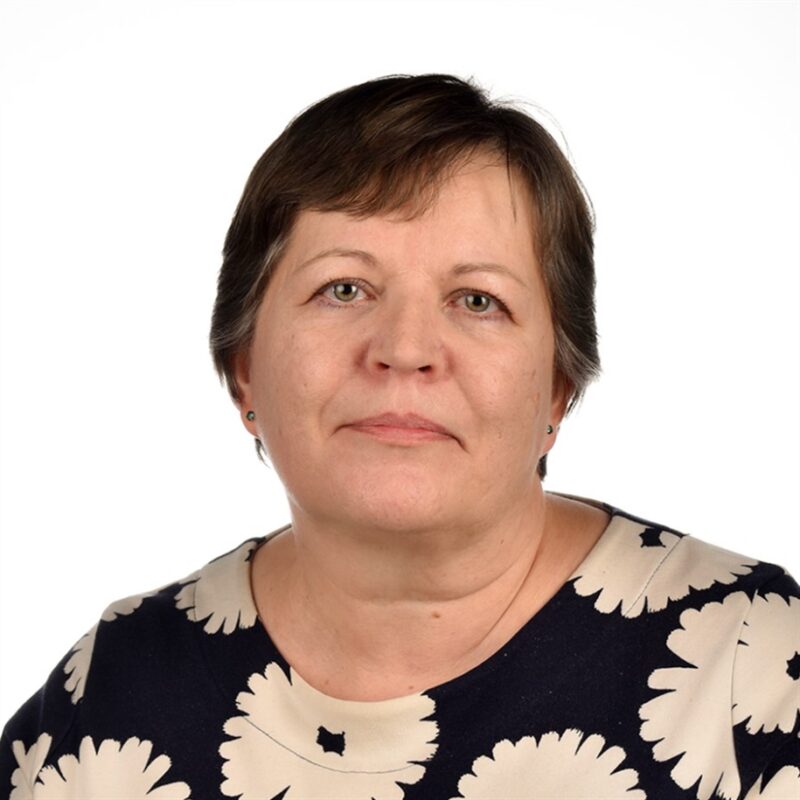 Lyhythiuksisella henkilöllä on yllään tummansininen paita, jossa on valkoisia kukkakuvioita. He katsovat suoraan kameraan tavallista valkoista taustaa vasten.