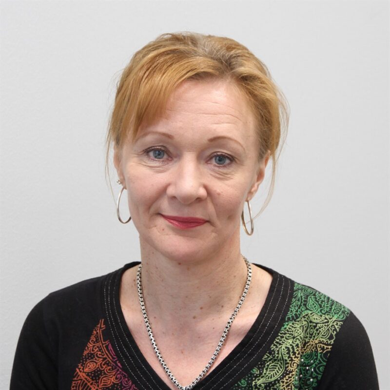 Vaaleanruskeat hiukset naisella on korvakorut ja musta toppi, jossa on värikkäitä kuvioita. Hän katsoo suoraan kameraan tavallista vaaleanharmaata taustaa vasten.