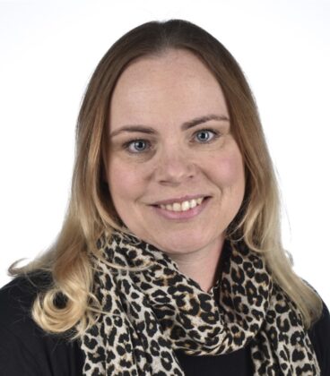 Nainen, jolla on vaaleanruskeat hiukset ja siniset silmät, hymyilee kameralle, yllään musta toppi ja leopardikuvioinen huivi tavallista valkoista taustaa vasten.