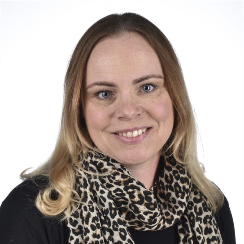 Nainen, jolla on vaaleanruskeat hiukset ja siniset silmät, hymyilee kameralle, yllään musta toppi ja leopardikuvioinen huivi tavallista valkoista taustaa vasten.