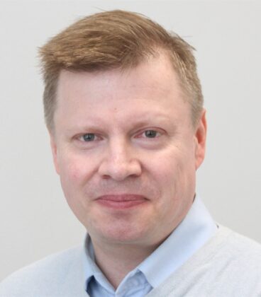 Miehellä, jolla on lyhyet, vaaleanruskeat hiukset, on yllään vaaleansininen kauluspaita ja harmaa pusero. Hän katsoo kameraan neutraalilla ilmeellä yksinkertaista, vaaleaa taustaa vasten.