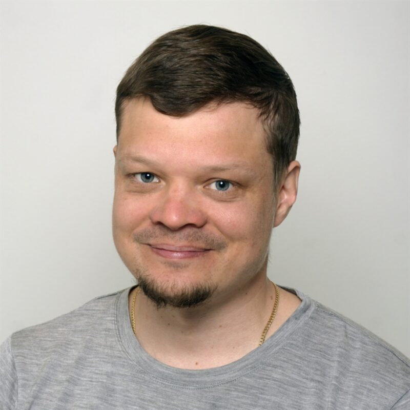 Mies, jolla on lyhyet ruskeat hiukset ja vuohenparkki hymyilee kameralle. Hänellä on yllään harmaa t-paita ja kultainen ketjukaulakoru. Tausta on tasainen ja vaalea.