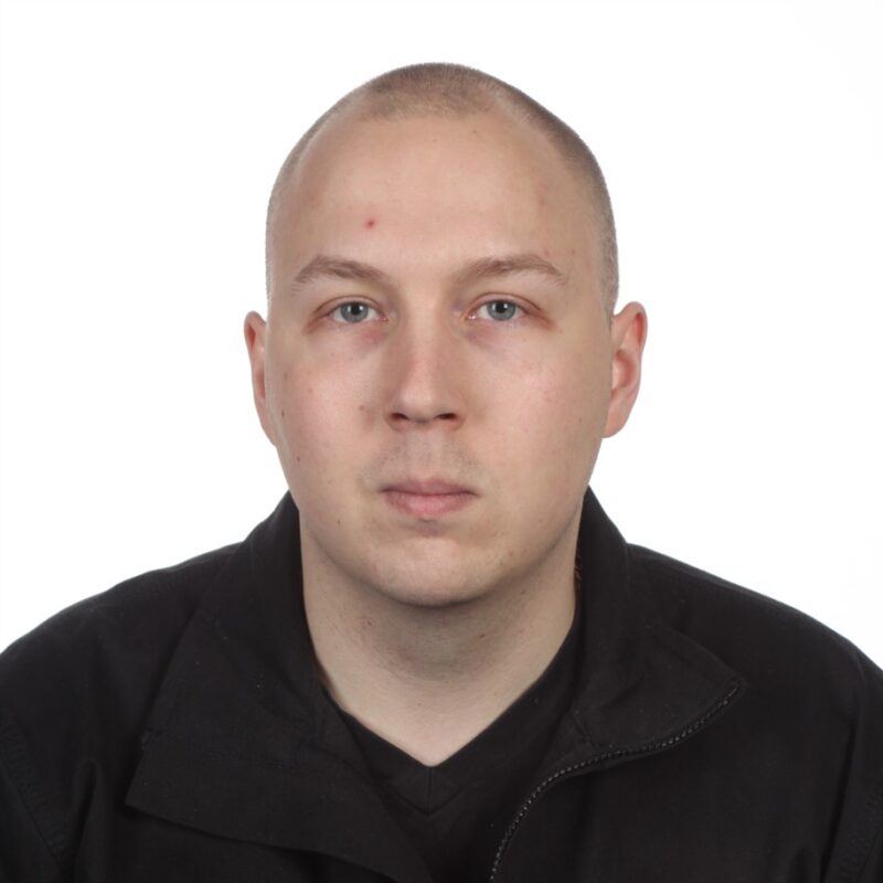 Henkilö, jolla on ajeltu pää ja jolla on yllään musta takki ja paita, katsoo suoraan kameraan valkoista taustaa vasten.