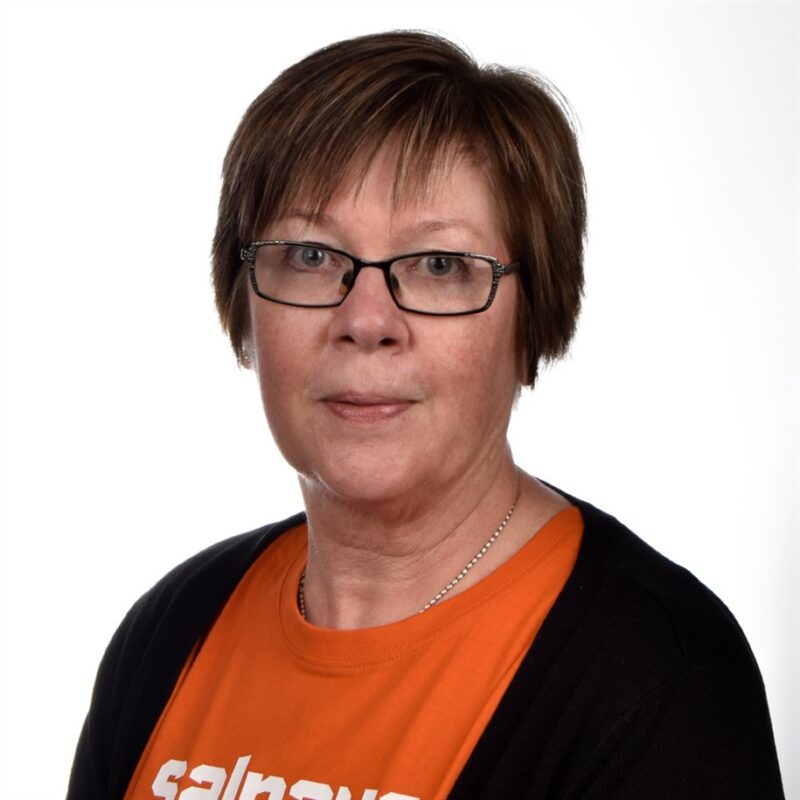 Lyhyet ruskeat hiukset ja silmälasit omaava henkilö on pukeutunut oranssiin paitaan ja mustaan takkiin tavallista valkoista taustaa vasten.